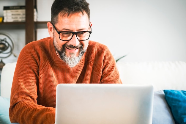 Homem de negócios bonito de meia-idade está usando um laptop e sorrindo enquanto trabalha em casa Homens bonitos maduros trabalhando em notebook de computador na sala de estar sentados no sofá e sorrindo na tela