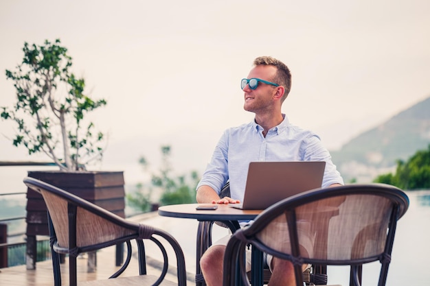 Homem de negócios bonito e bem-sucedido em óculos de sol trabalha em um laptop sentado perto da piscina Trabalho remoto Freelancer