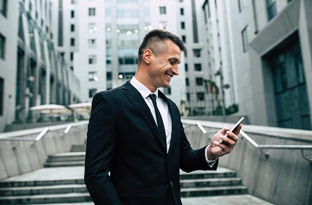 Homem de negócios bonito e confiante moderno inteligente em uma roupa formal usa um telefone inteligente ao ar livre