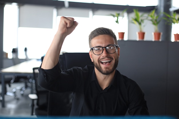 Homem de negócios bonito está mantendo o braço levantado e expressando alegria no escritório.