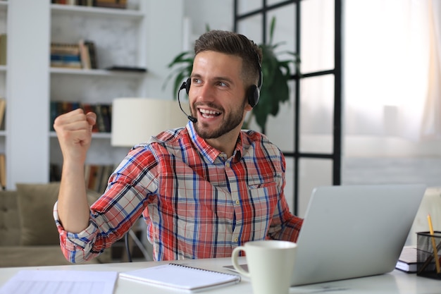 Homem de negócios bonito está mantendo os braços levantados e expressando alegria no escritório em casa.