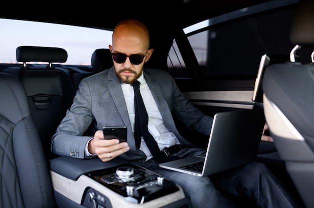 Homem de negócios bonito usando seu telefone celular em um carro moderno com um motorista no centro da cidade conceito de sucesso empresarial viajando de luxo