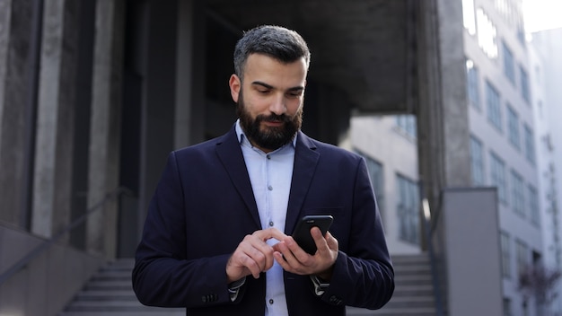 Homem de negócios bonito usando smartphone em pé perto do centro de negócios.