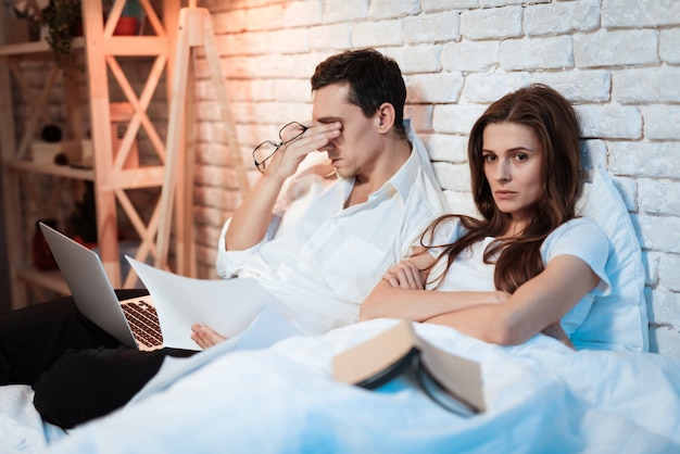 Homem de negócios cansado que senta-se na cama que trabalha no portátil.