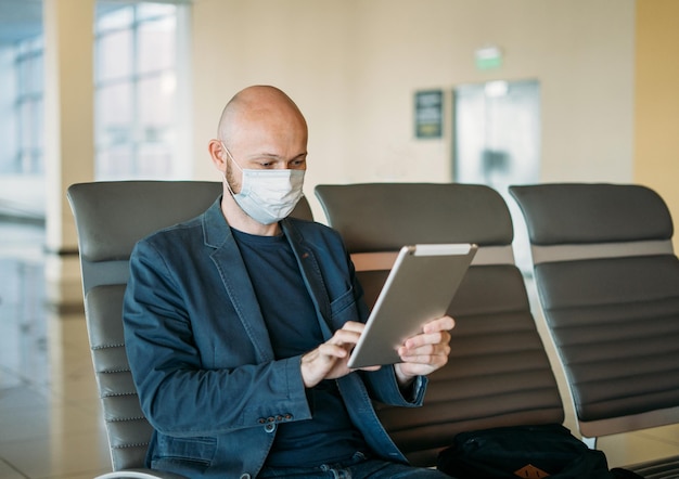 Homem de negócios careca com máscara facial médica usando tablet no aeroporto. Conceito de quarentena de coronavírus