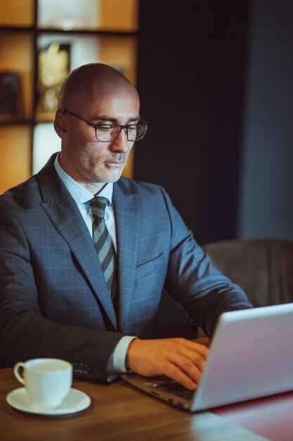 Homem de negócios careca de meia idade em suíte trabalhando no laptop sentado no escritório moderno. Bonito ousado