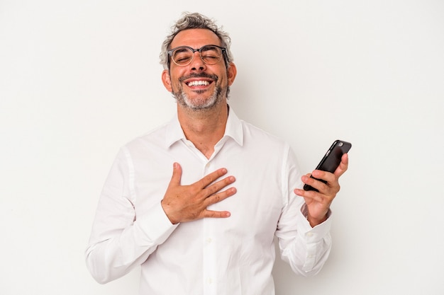 Homem de negócios caucasiano de meia-idade segurando um telefone celular isolado no fundo branco ri alto, mantendo a mão no peito.