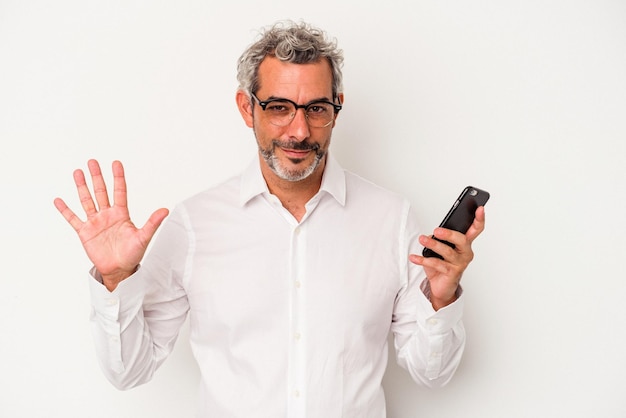 Homem de negócios caucasiano de meia-idade segurando um telefone celular isolado no fundo branco sorrindo alegre mostrando o número cinco com os dedos