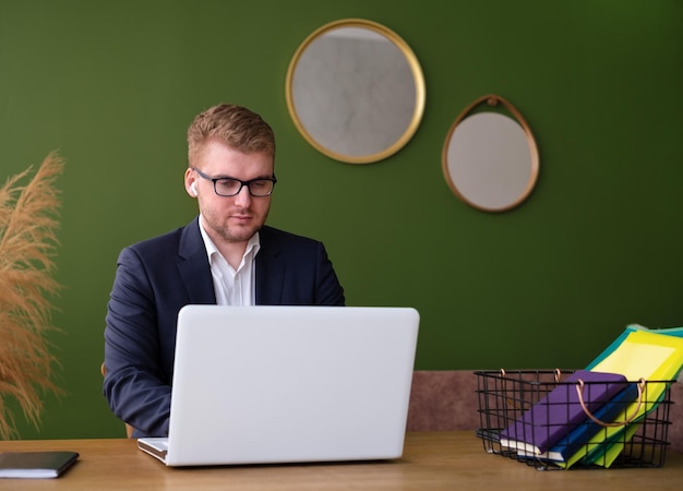 Homem de negócios caucasiano loiro usando óculos com fones de ouvido trabalha com laptop branco no escritório em casa