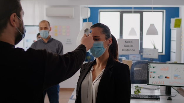 Homem de negócios com máscara protetora, verificando a temperatura dos colegas usando termômetro infravermelho para evitar a infecção do vírus. colegas de trabalho mantendo distância social para prevenir a propagação do coronavírus