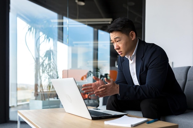 Homem de negócios concentrado conversando com colegas em videochamada usando laptop sentado no escritório homem asiático trabalhando on-line