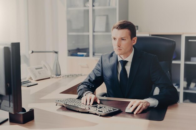 Homem de negócios concentrado sério senta-se na frente do computador no local de trabalho teclados informações veste terno prepara projeto de negócios para o próximo ano posa em armário espaçoso Pessoas e finanças