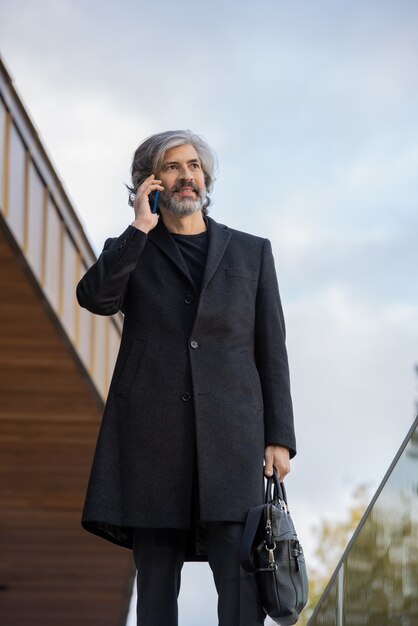 Foto homem de negócios confiante de cabelos grisalhos vestindo um casaco preto usando smartphone enquanto consulta um cliente ou faz uma consulta no exterior do centro de negócios