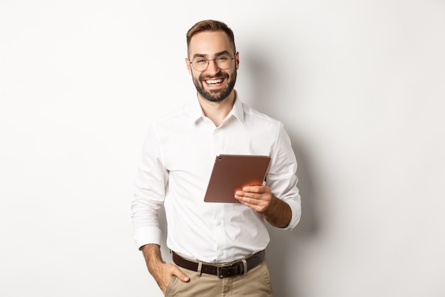 Homem de negócios confiante segurando o tablet digital e sorrindo, de pé contra um fundo branco.