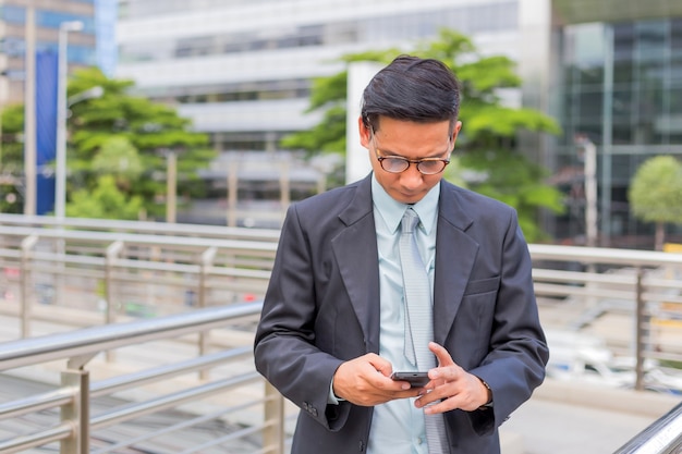 Homem de negócios considerável novo de Ásia com seu smartphone que está na passagem da cidade moderna.