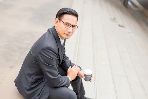 Homem de negócios considerável novo de Ásia que bebe o café quente na cidade moderna. Quebrando e relaxando o conceito de negócio
