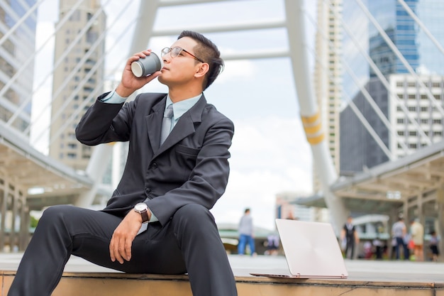 Homem de negócios considerável novo de Ásia que bebe o café quente na cidade moderna. Quebrando e relaxando o conceito de negócio