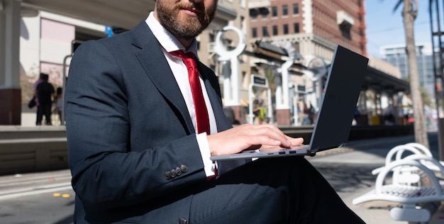 Homem de negócios cortado de terno sentado no banco na cidade ao ar livre trabalhando on-line em blogs de negócios de laptop