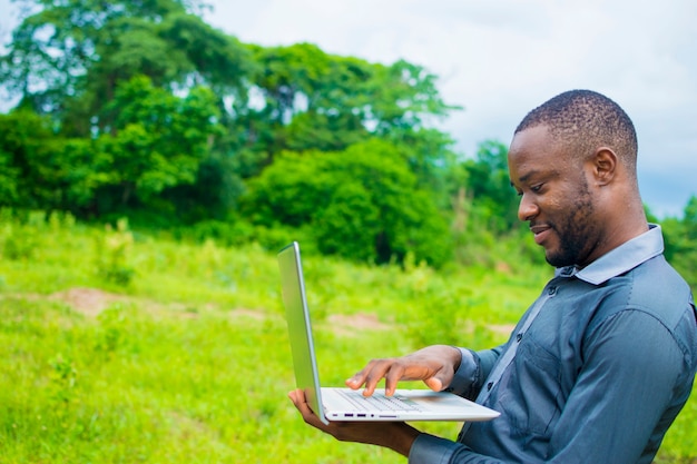 Homem de negócios da áfrica negra bonito jovem de pé em uma fazenda usando seu laptop.