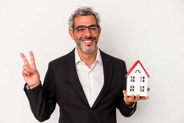 Homem de negócios de meia idade segurando contas e modelo de casa isolado em fundo azul alegre e despreocupado, mostrando um símbolo de paz com os dedos