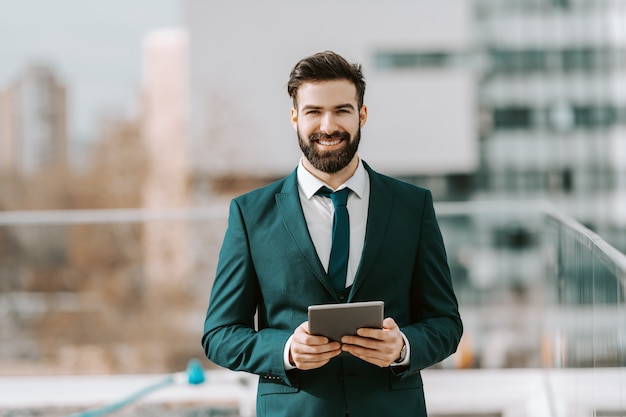 Homem de negócios de sorriso no vestuário formal usando a tabuleta ao estar no telhado.