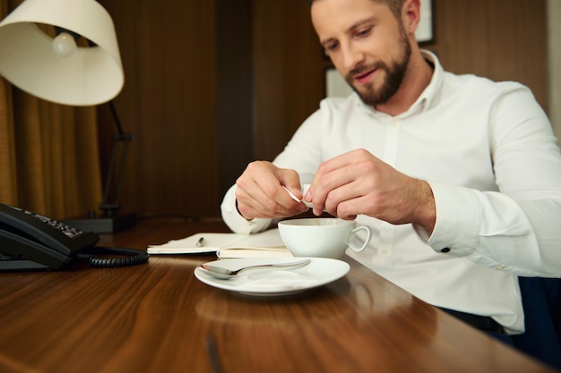 Homem de negócios descontraído e bonito em uma camisa branca polvilhando açúcar em palitos em um copo de cerâmica com uma bebida de café acabado de fazer, sentado a uma mesa de madeira e desfrutando de uma pausa para o café no quarto de hotel