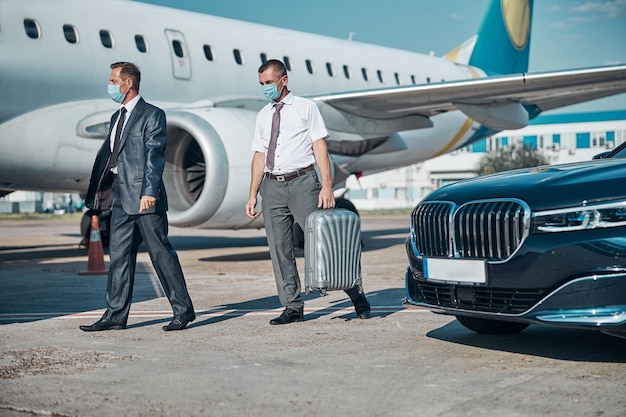 Homem de negócios elegante com assistente com mala está vindo de carro para a pista do aeroporto para voar durante a quarentena