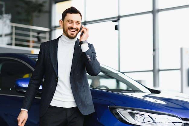 Homem de negócios elegante com o telefone na mão em pé perto do novo carro azul