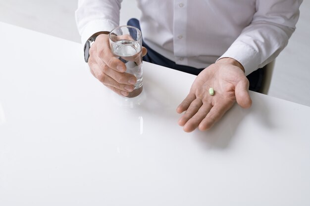 Homem de negócios elegante de camisa branca segurando um copo de água e analgésico enquanto está sentado à mesa e vai tomar o comprimido