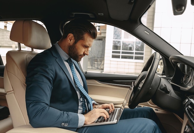 Homem de negócios elegante de terno digitando algo no laptop no carro