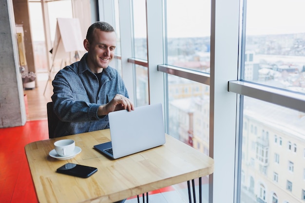 Homem de negócios feliz bem sucedido está sentado em uma mesa em um café segurando uma xícara de café e usando um laptop