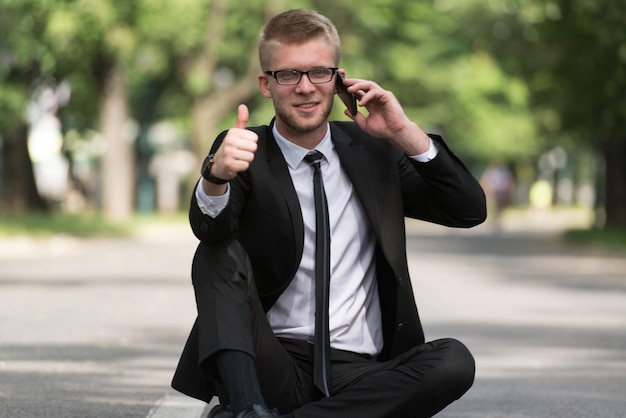 Homem de negócios feliz usando telefone celular fora sentado no asfalto