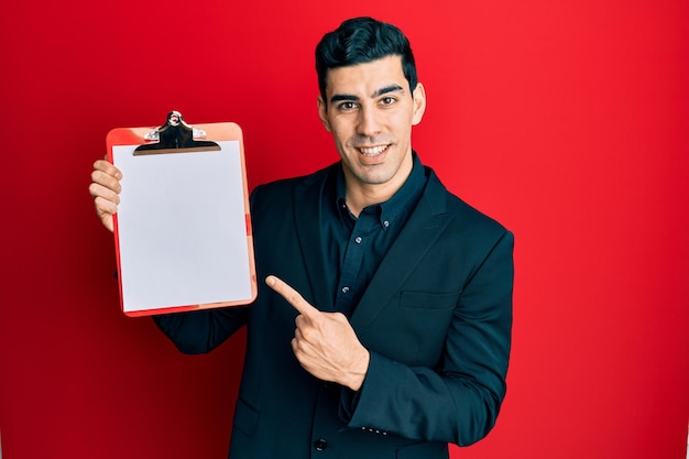Foto homem de negócios hispânico bonito segurando a área de transferência com espaço em branco sorrindo feliz apontando com a mão e o dedo
