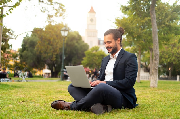Homem de negócios hispânico bonito sentado em posição de lótus no parque público usa laptop