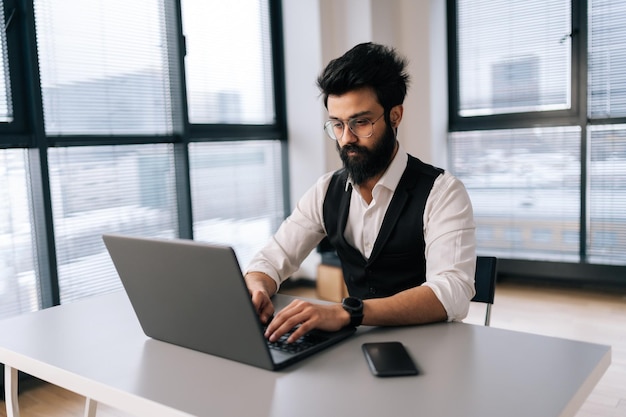 Homem de negócios indiano barbudo em trajes formais trabalhando digitando no laptop sentado na mesa no escritório de coworking leve no fundo da janela