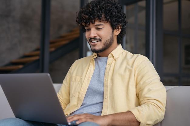 Homem de negócios indiano sorridente usando computador portátil assistindo cursos de treinamento sentado no escritório moderno