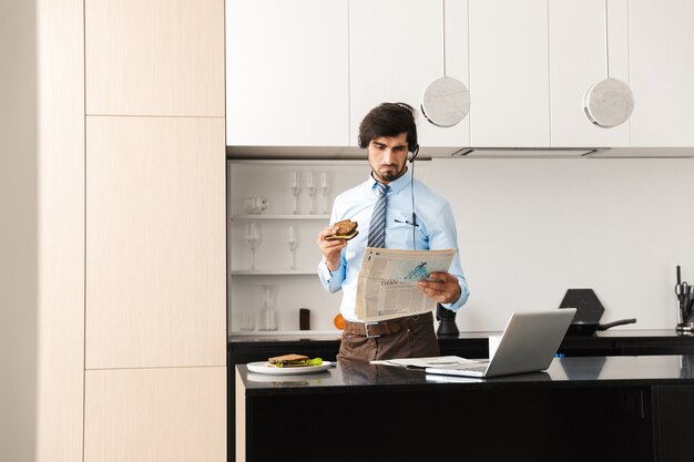 Homem de negócios jovem bonito na cozinha comer sanduíche usando o computador portátil com fones de ouvido, lendo o jornal.