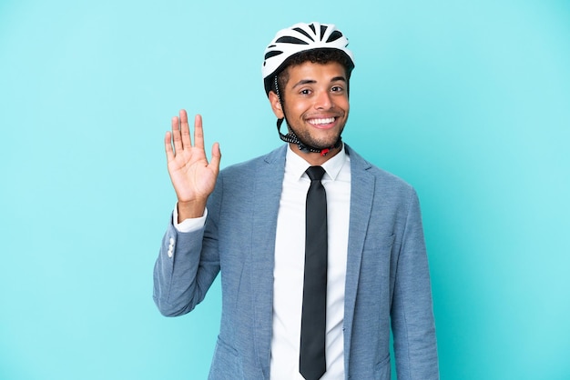 Homem de negócios jovem brasileiro com capacete de bicicleta isolado em fundo azul saudando com a mão com expressão feliz