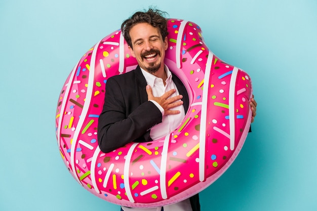 Homem de negócios jovem com donut inflável isolado sobre fundo azul ri alto, mantendo a mão no peito.