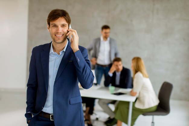 Homem de negócios jovem e bonito parado confiante no escritório, em frente à sua equipe, usando o telefone celular