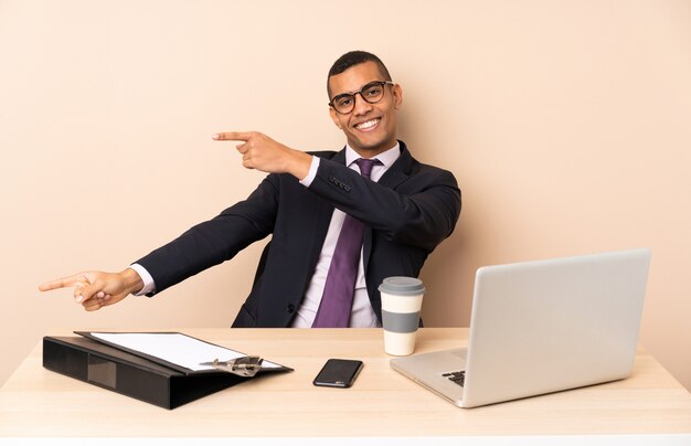 Homem de negócios jovem em seu escritório com um laptop e outros documentos, apontando o dedo para o lado e apresentando um produto