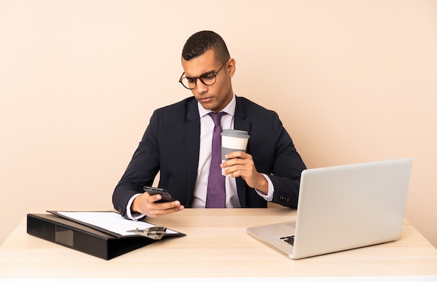 Homem de negócios jovem em seu escritório com um laptop e outros documentos segurando café para levar e um celular