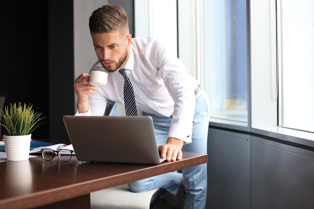 Homem de negócios jovem pensativo está usando o computador e bebendo café enquanto trabalhava no escritório.