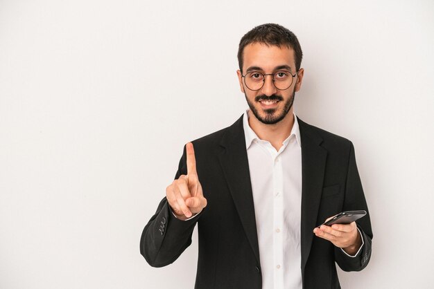 Homem de negócios jovem segurando um telefone celular isolado no fundo branco, mostrando o número um com o dedo