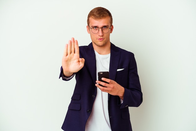 Homem de negócios jovem segurando um telefone móvel isolado no branco em pé com a mão estendida, mostrando o sinal de pare, impedindo-o.