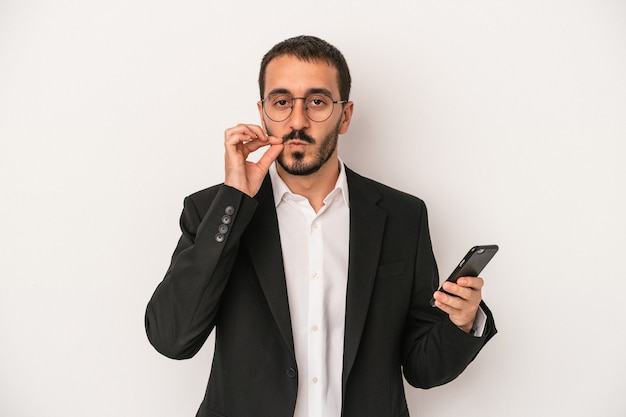 Homem de negócios jovem segurando um telefone móvel isolado no fundo branco com os dedos nos lábios, mantendo um segredo.