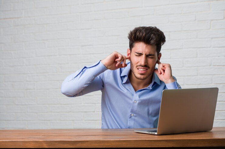 Foto homem de negócios jovem sentado e trabalhando em um laptop cobrindo as orelhas com as mãos