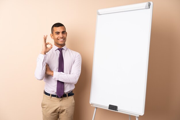 Homem de negócios jovem sobre parede isolada, dando uma apresentação no quadro branco e mostrando sinal okey com os dedos