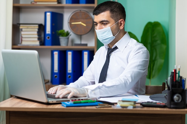 Homem de negócios jovem trabalhando no laptop com luvas e máscara protetora médica preta