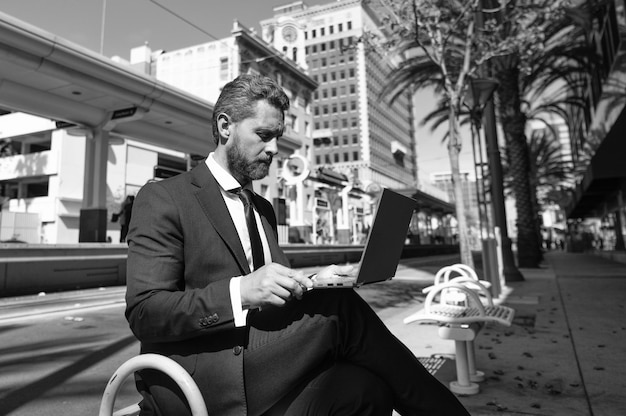 Homem de negócios maduro de terno sentado no banco na cidade ao ar livre trabalhando on-line na educação do laptop